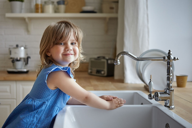 Charming little girl in blue dress washing hands in kitchen. Cute female kid looking and smiling at camera, helping mother, doing dishes, standing at sink. Kids, childhood, cooking and housework