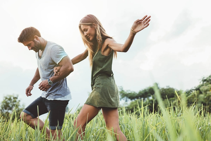 Outdoor shot of young couple walking through meadow hand in hand. Man and woman talking walk through grass field in countryside.